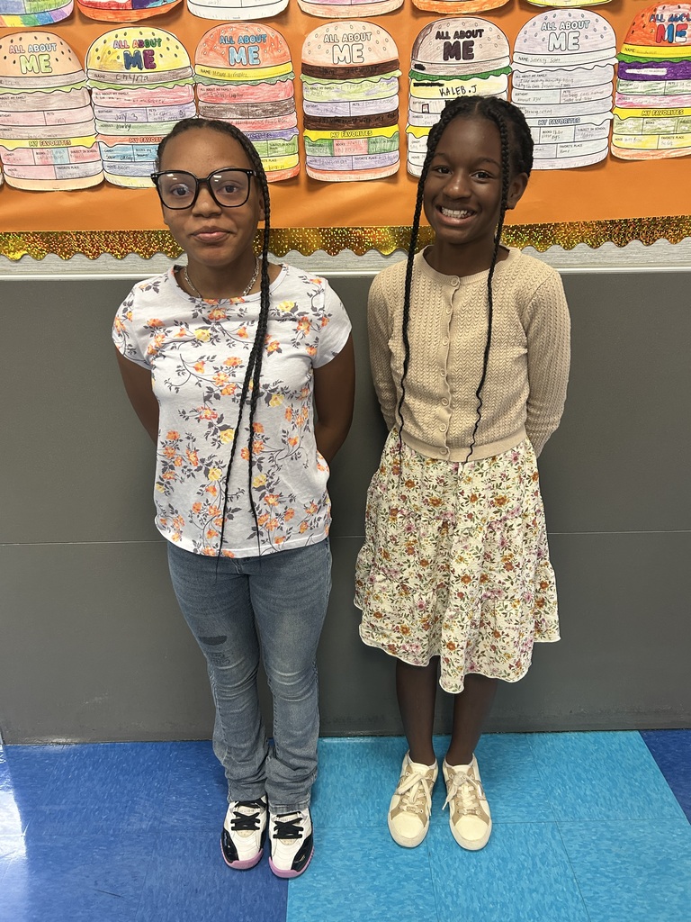 Two girls stand in front of a classroom wall with an orange backdrop, smiling. One wears glasses, a floral shirt, and jeans; the other has a beige sweater and floral skirt.