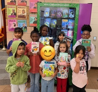 Children, holding books, stand in a group. Behind them, a bright book vending machine is displayed.