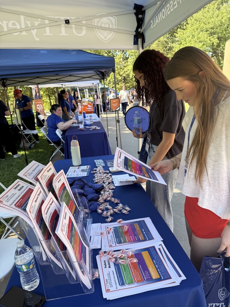 Two women at a booth, examining papers and candy. Tables covered in blue cloths. Blue tent with logo.