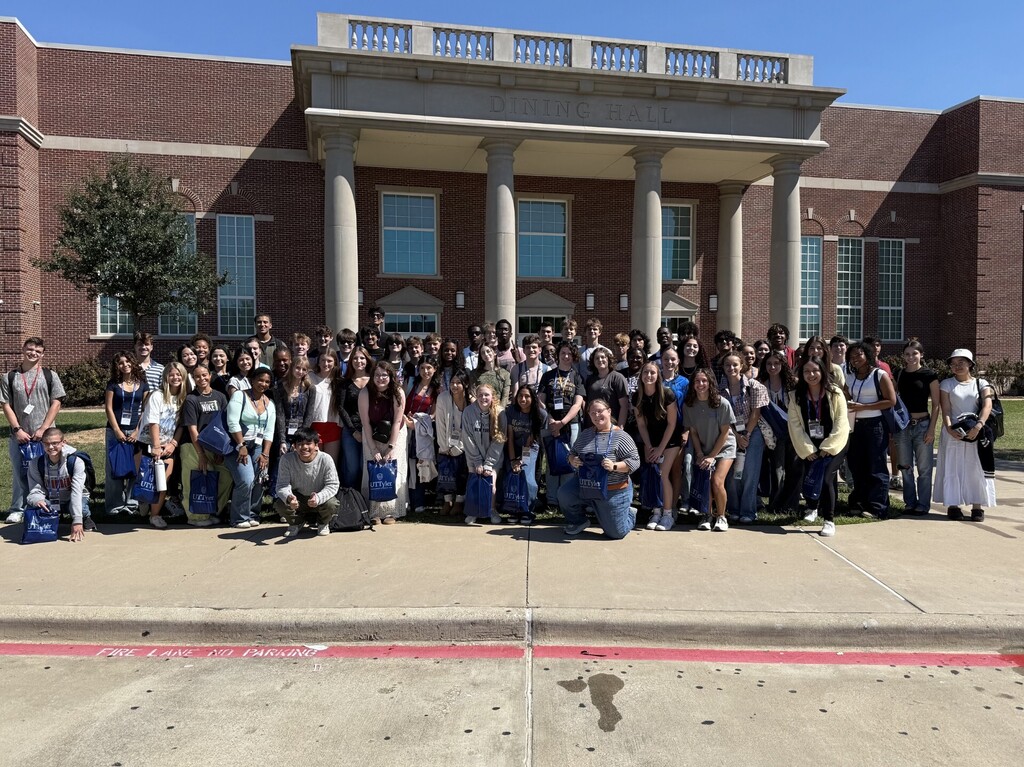 A group of students poses in front of a large building with a tree and a paved walkway.