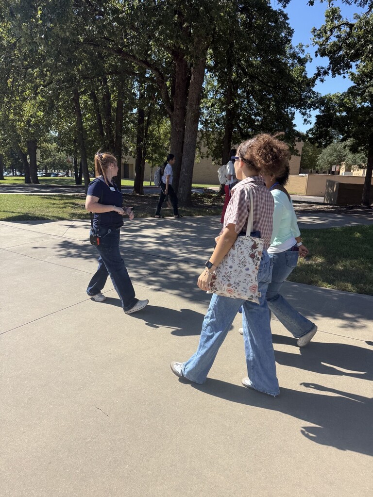 Three people walk in a line on a concrete pathway. One person carries a bag. Trees and grass are in the background.