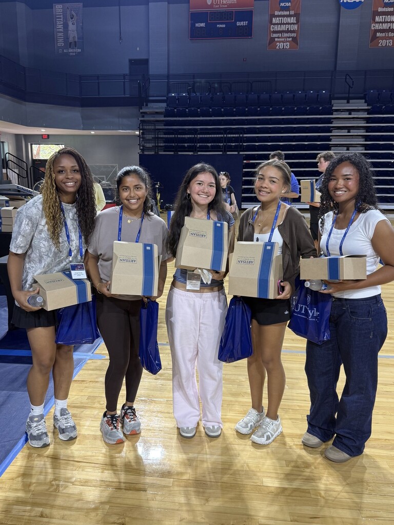 Five women stand on a basketball court, holding boxes and smiling. Two banners hang on the wall behind them.