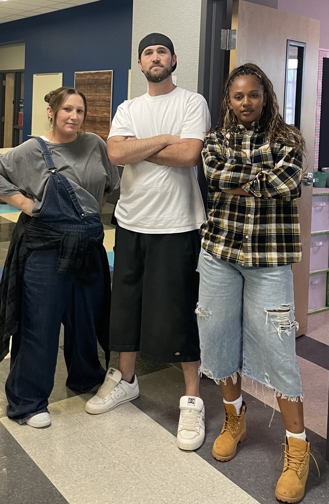 Three people stand together in a hallway. One wears a white shirt, another has a checkered shirt, and the third wears overalls.