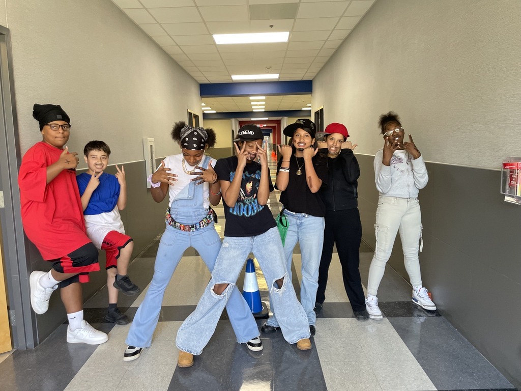 A group of seven young people in a hallway, standing and posing for a photo. Some wear hats and sneakers.