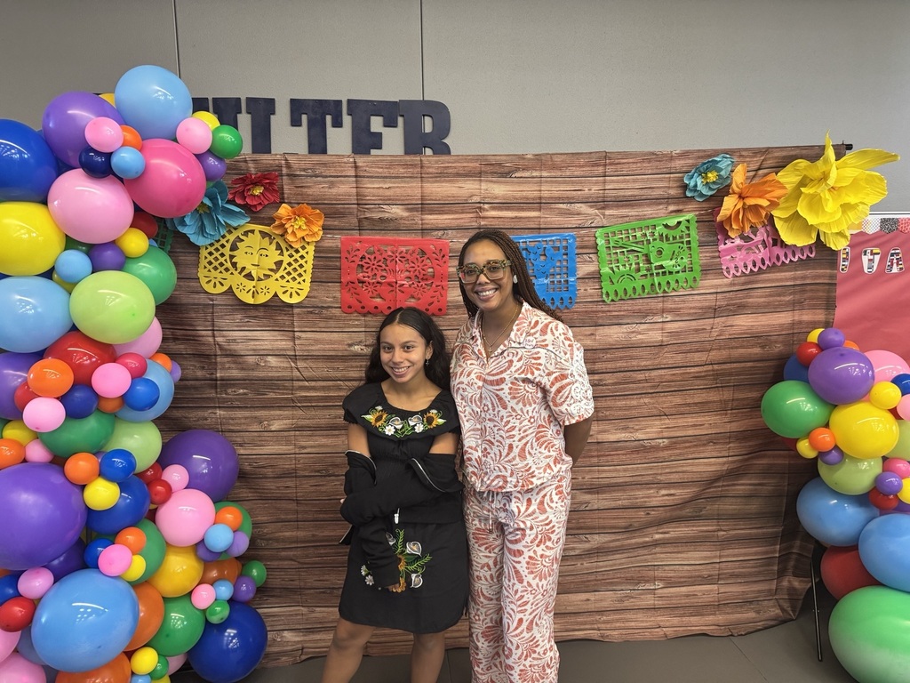 Two women stand in front of a wooden backdrop with a colorful balloon arch and decorations.