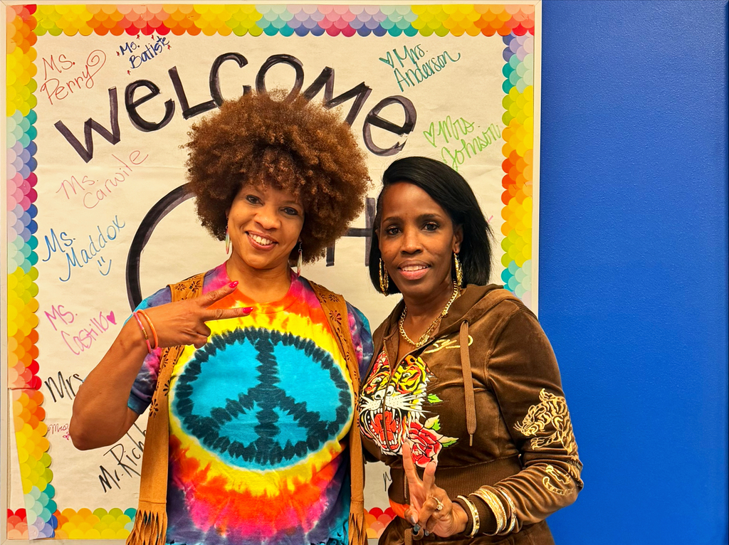 Two women pose before a vibrant, multicolored bulletin board with the word "Welcome" in large letters.