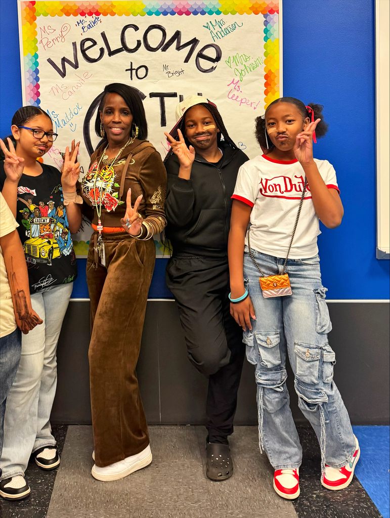 Four individuals pose for a photo in front of a "Welcome to T" banner. Two women and two girls are making peace signs.