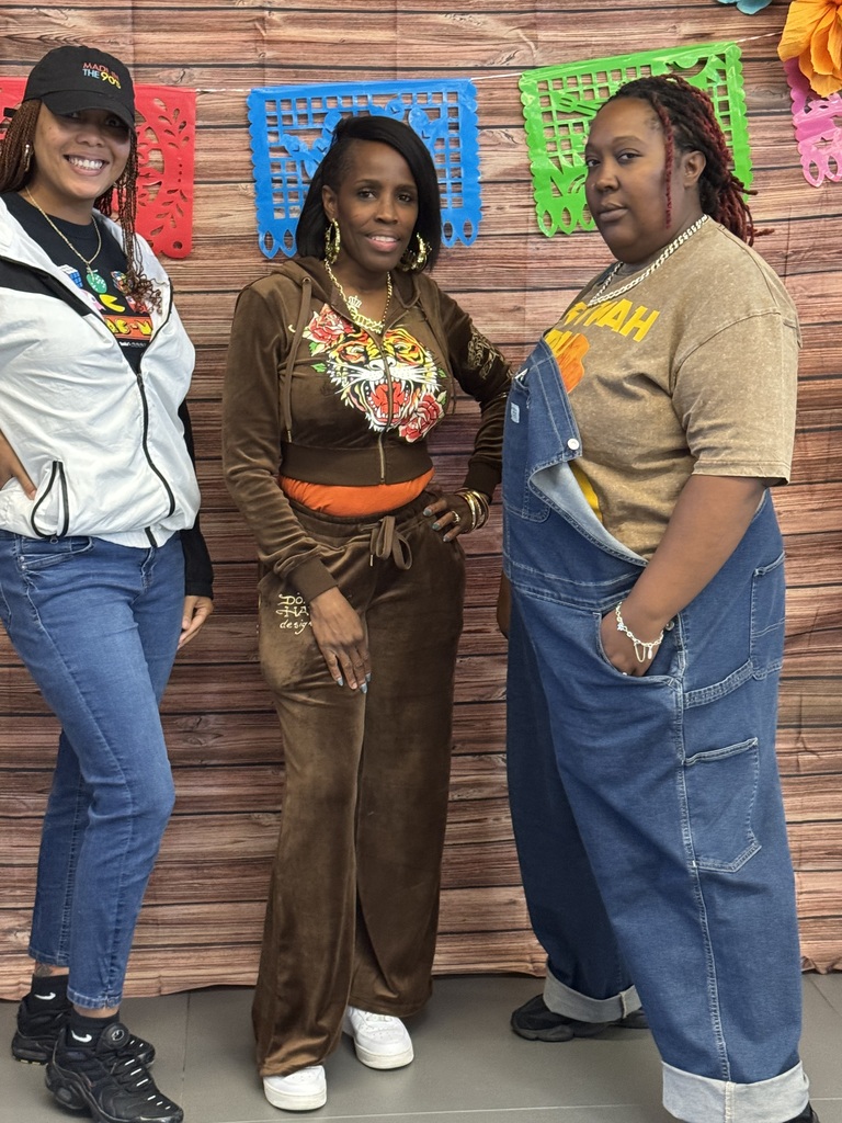 Three women stand together in front of a wooden backdrop. Each wears different colored tops and pants, with one in denim overalls.