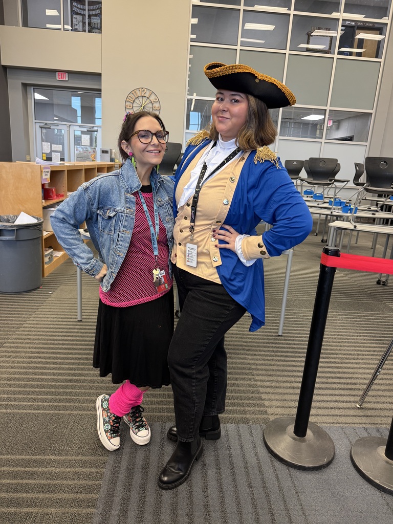 Two women stand in a room with a backdrop of chairs and tables. One wears glasses and a denim jacket, while the other dons a pirate hat and a blue coat.