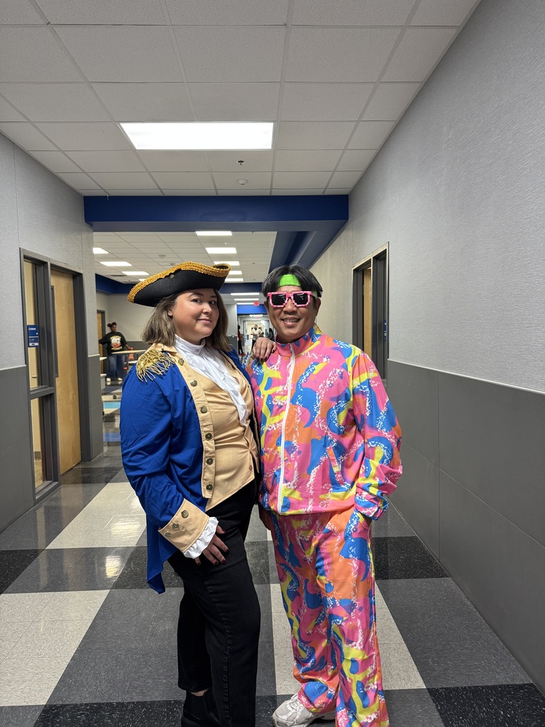 Two individuals dressed in vibrant costumes standing in a hallway with checkered floor tiles and white walls.