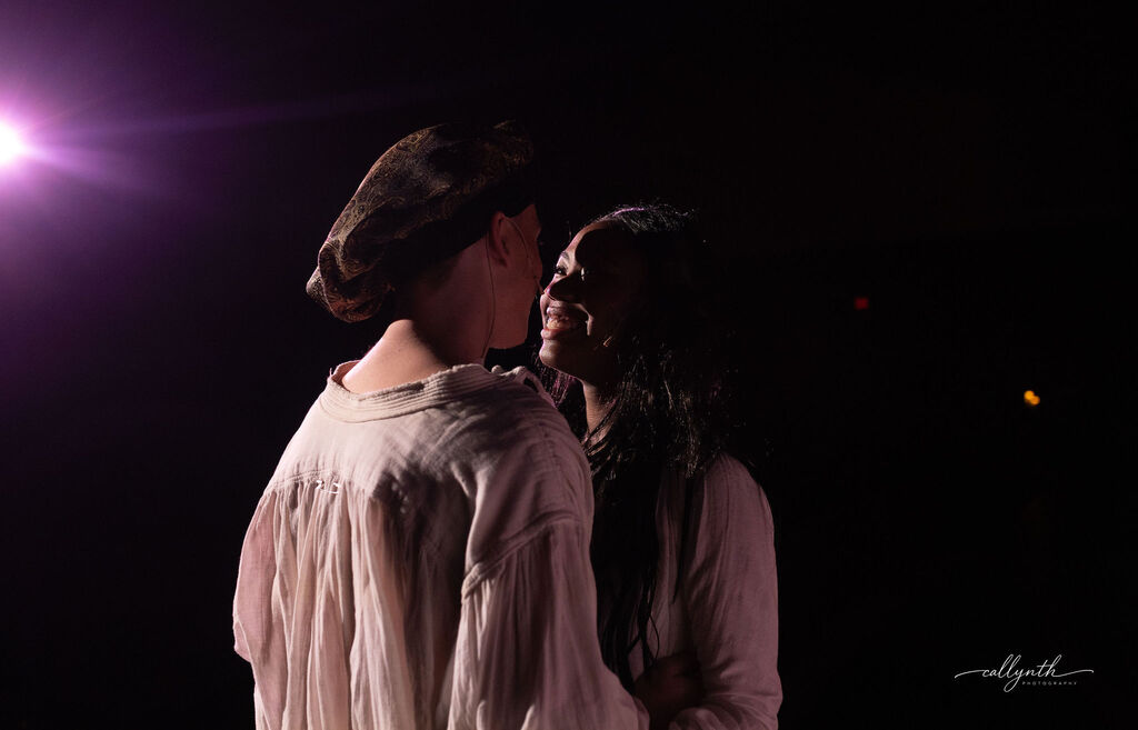 Two people in a dark setting, facing each other and looking close. They are illuminated by a bright light above.