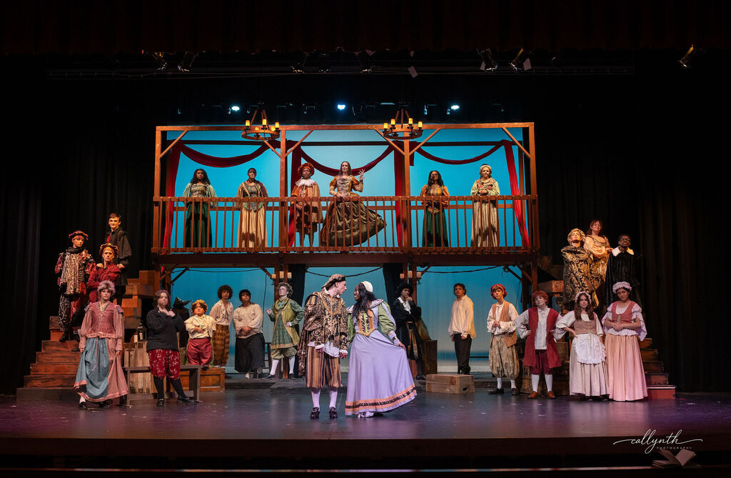 Stage with people in costume. Blue background with two levels of people. A group in front, others on balcony.