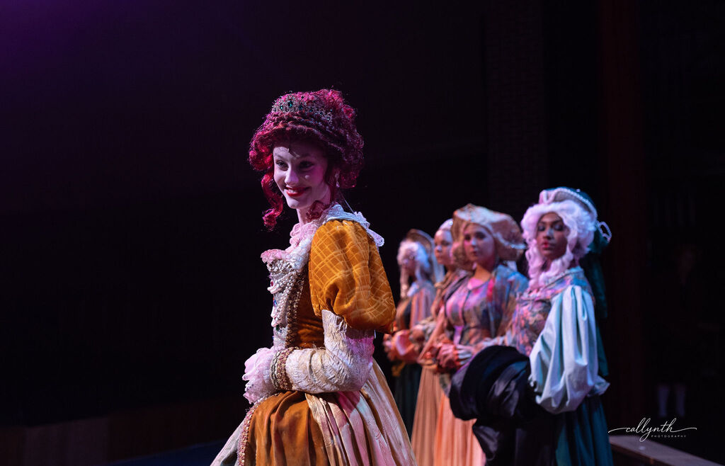 Five women, one in a red wig and elaborate dress, on stage with purple lighting.