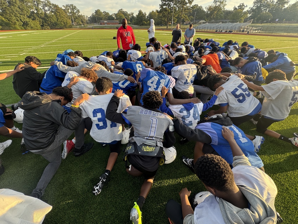A football team huddles together on a grassy field, wearing blue and white jerseys, with numbers visible.