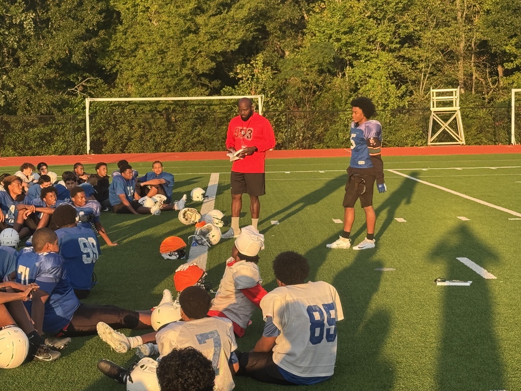 Group of players in blue and white uniforms sit on a field with a coach in red.