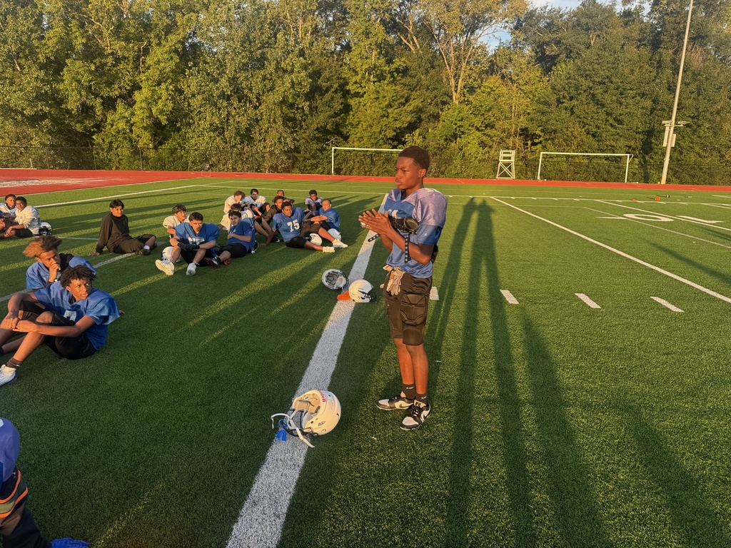 On a field, a coach speaks to students wearing jerseys, some seated and others standing. The field is marked with white lines. Trees are in the background.