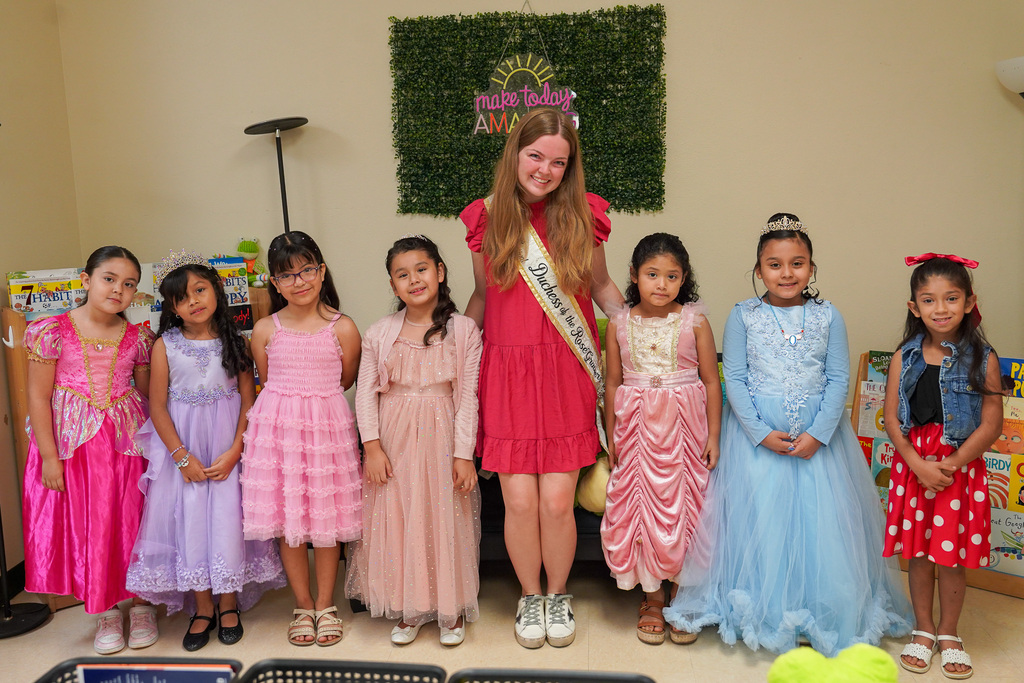 Several girls in dresses stand together, one with a sash. A woman stands in the middle.