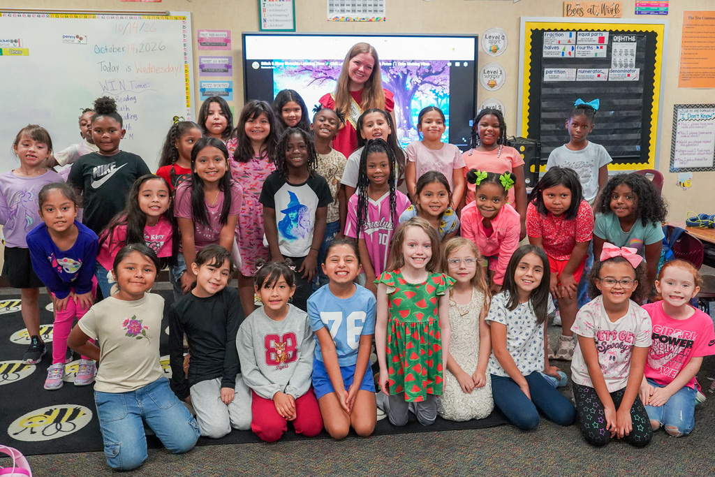 Classroom photo with children in various clothing, sitting or standing, in front of a whiteboard and classroom materials.