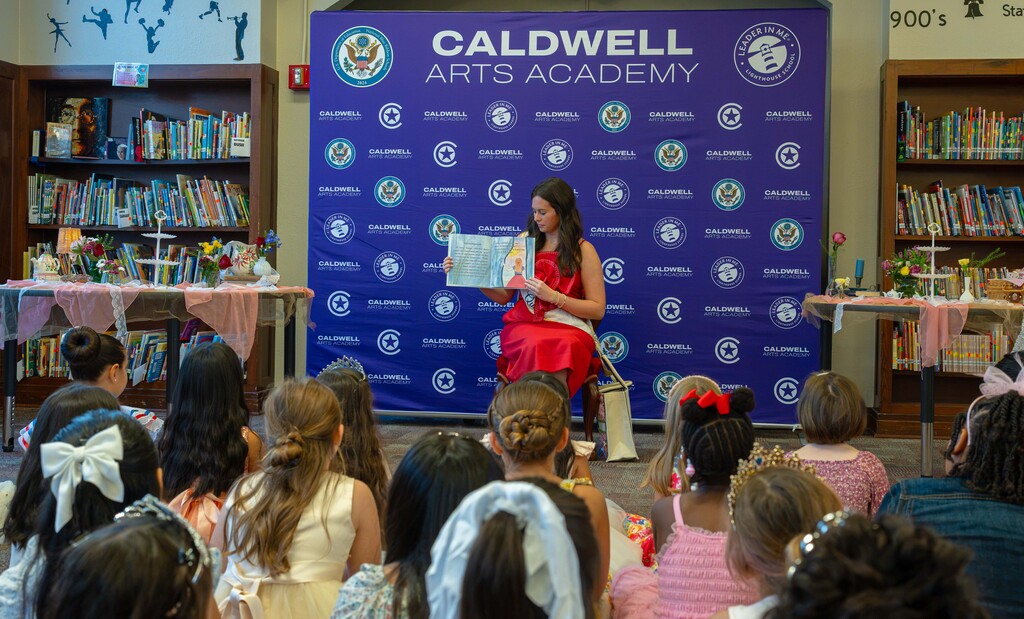 A woman in a red dress reads a book to a group of children in a library.