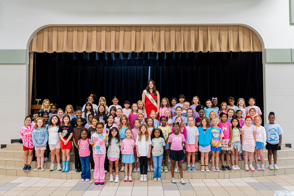 A woman stands in front of a group of children on a stage, all wearing various colors and styles.