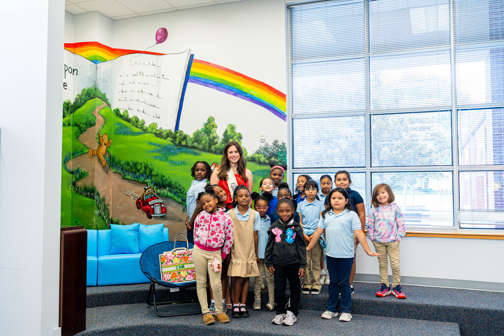 Children stand on a stage in a room with a rainbow and landscape mural behind them.