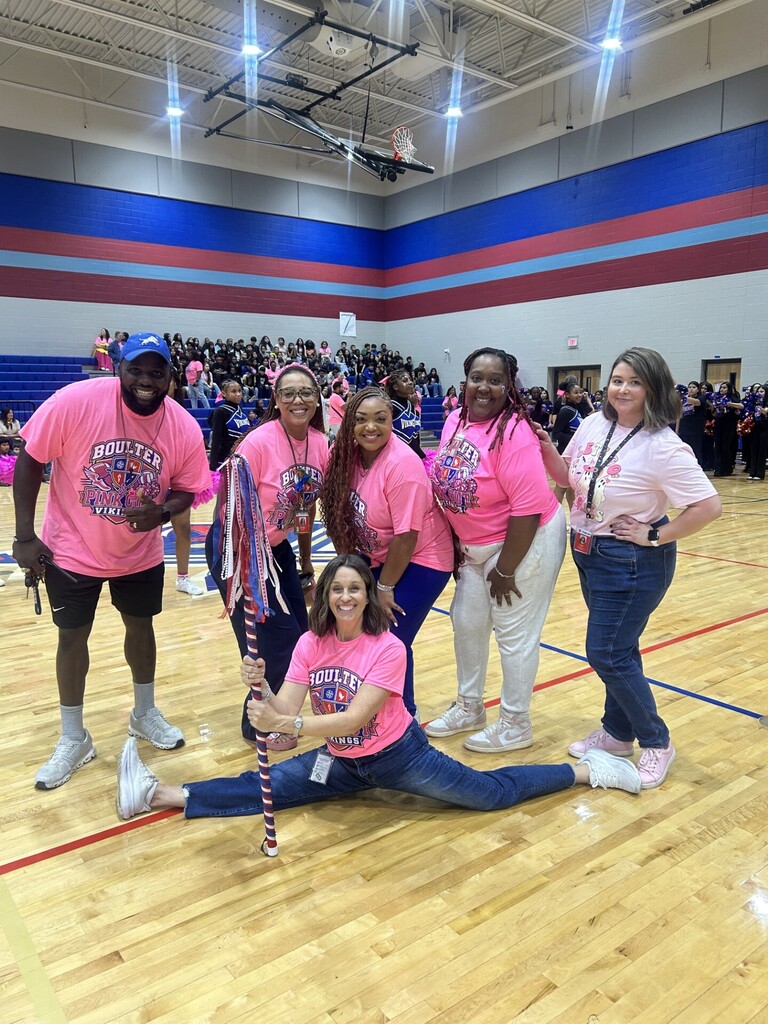 A group of women and a man in pink shirts and sneakers pose on a basketball court.