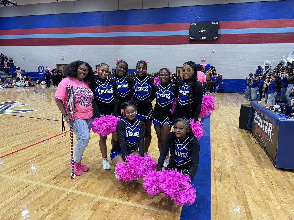 Girls in cheerleading uniforms pose with pom-poms on a basketball court, with a cheering crowd in the background.