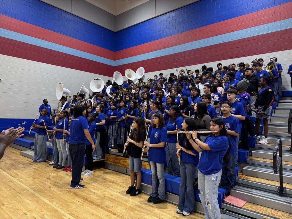 A band performs inside a gymnasium. Some hold instruments. Steps are behind them, and bleachers are visible.
