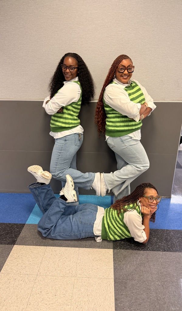 Three people dressed in matching green vests and jeans, two standing and one sitting on the floor.