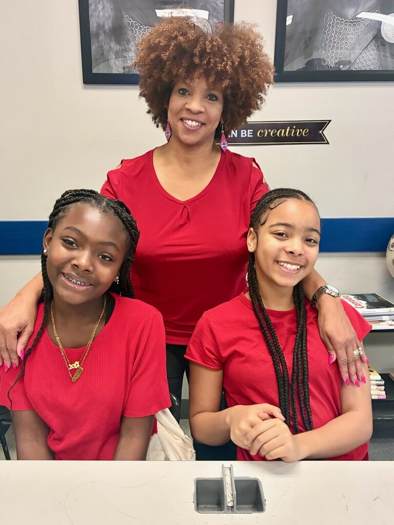 Woman with curly hair standing behind two girls in red shirts, all smiling. Behind them, two framed photos and a banner.