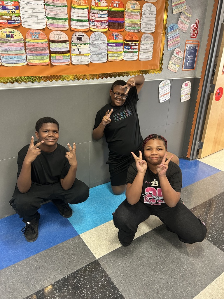 Three individuals in black shirts and pants, two making peace signs, are posing for a photo against a wall with posters and checkered floor.