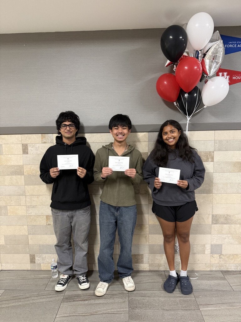 Three people stand holding certificates; one wears glasses, another a green shirt. Behind them, balloons in red, white, and black decorate the wall.