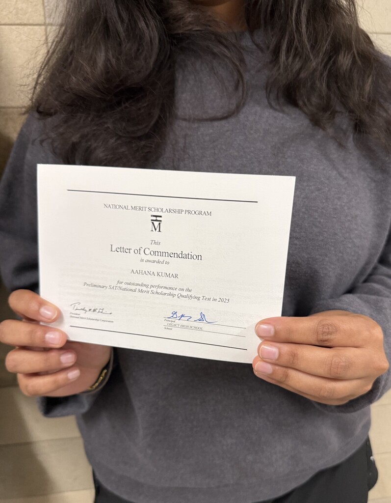 A person holds a white paper, "Letter of Commendation," with black text and a logo, wearing a gray top and black pants.