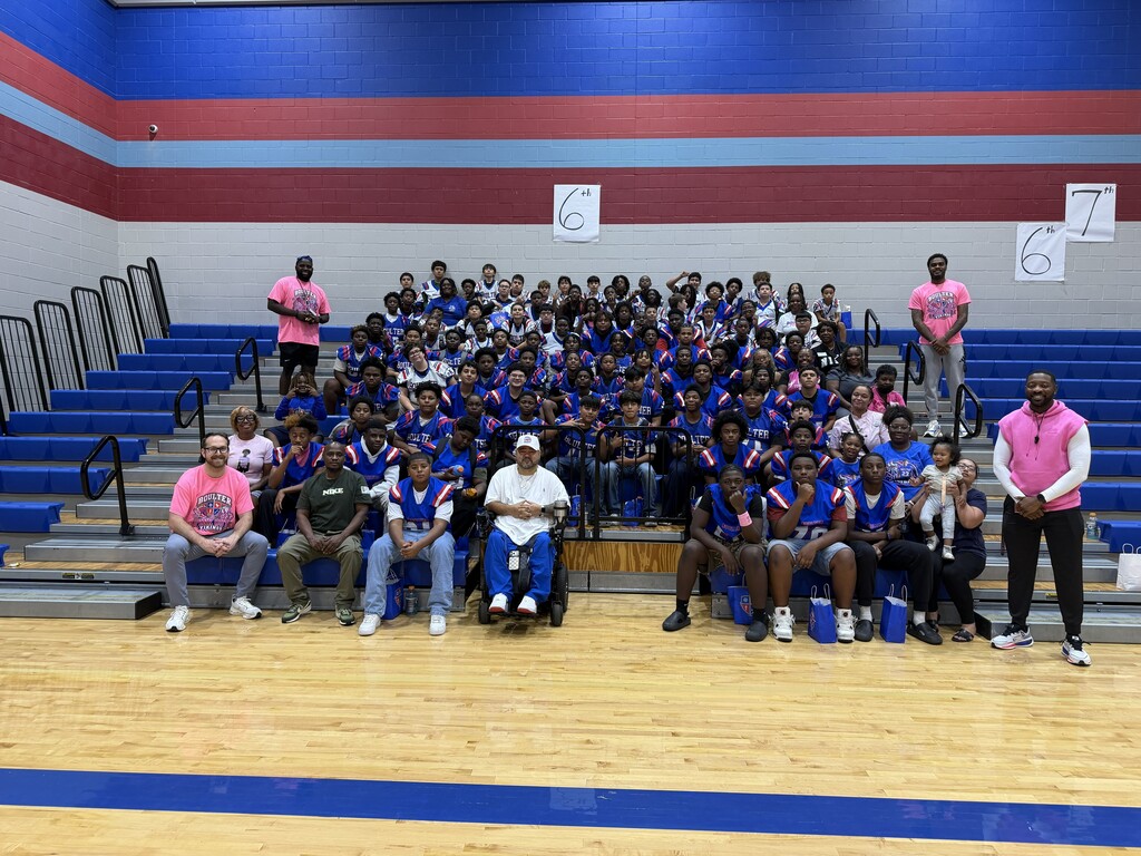 A sports team in blue jerseys and pink tops poses on a gym's bleachers. Some sit, others stand, and two adults in pink are on the right.