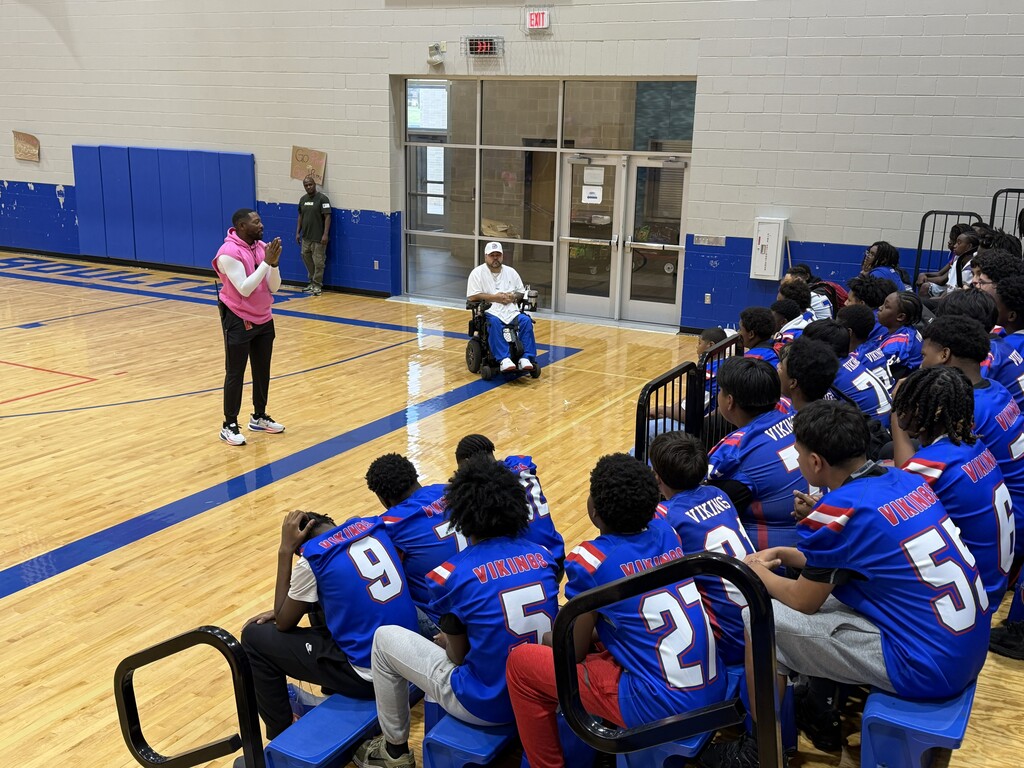 A coach in a pink shirt gives a speech to a team in blue uniforms sitting in chairs.