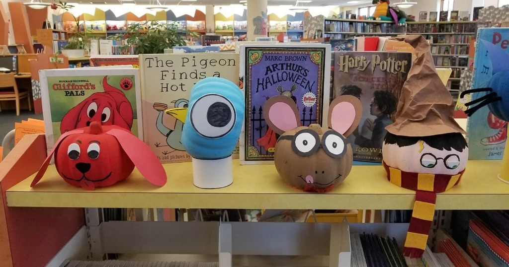 Four pumpkins that are decorated as Clifford, the Pigeon, Arthur, and Harry Potter are displayed on a bookshelf with their corresponding books behind them.