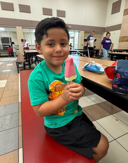 A boy smiles while holding a pink popsicle inside a cafeteria with tiled floors and tables.