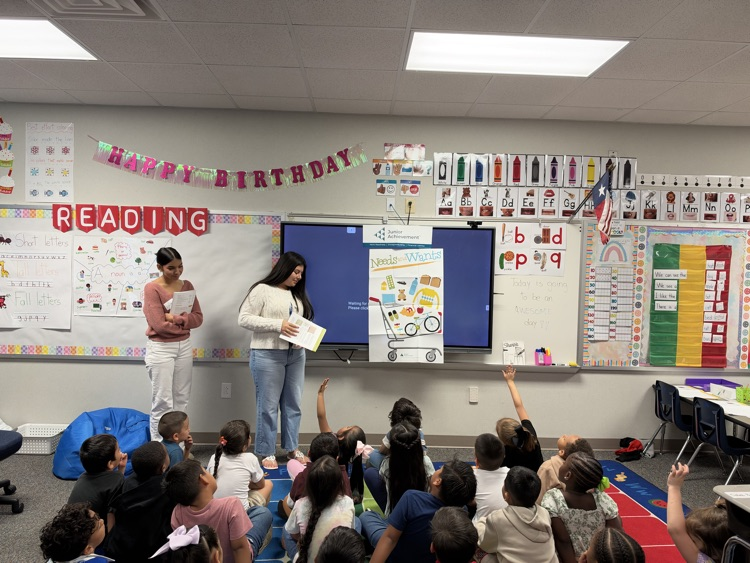 Two teachers and students in a classroom with a TV screen, chairs, and a rug.
