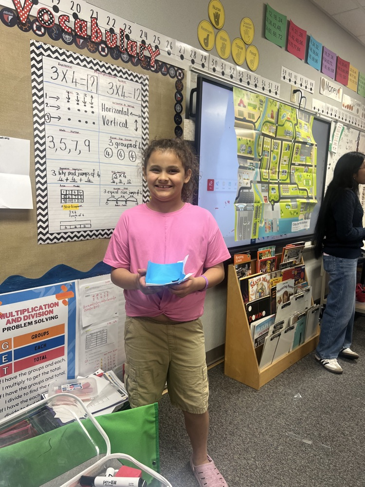 A girl holds paper in classroom with a teacher. A large board, books, and a shelf with colorful items behind her.