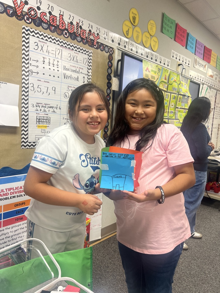 Two young girls holding colorful paper in a classroom with colorful bulletin boards and a woman nearby.