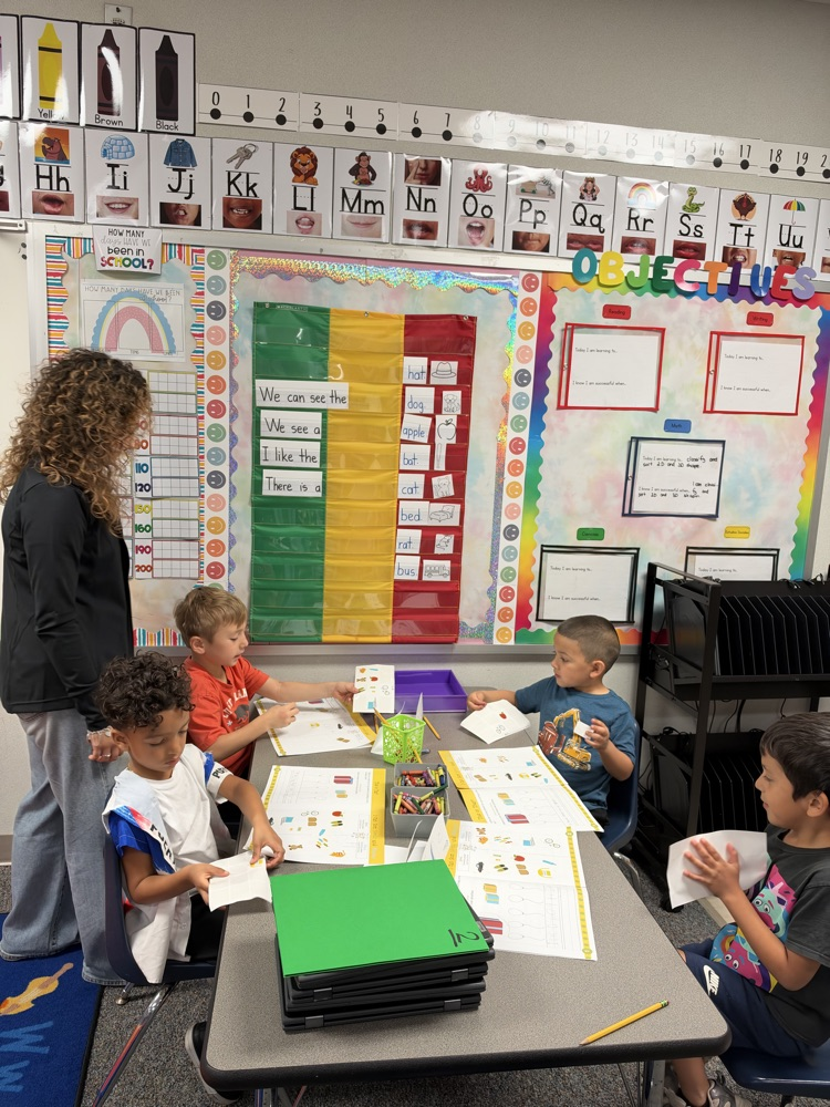 Teacher standing by children at a table. Paper, pencils, folders, and crayons on table.