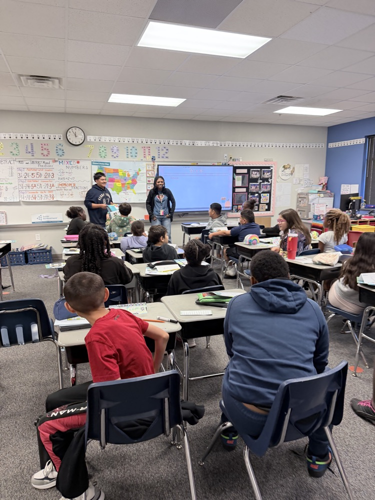 Students seated in chairs facing a screen in a classroom; two teachers standing at front of room.
