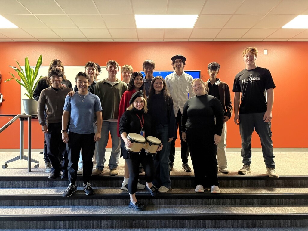 A group of students and a teacher stand on stairs with instruments, including drums. Behind them is a plant and an orange wall.