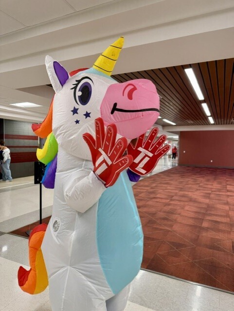 Person in inflatable unicorn costume with red gloves, hands raised in front of body. Brown carpet on floor, beige walls.