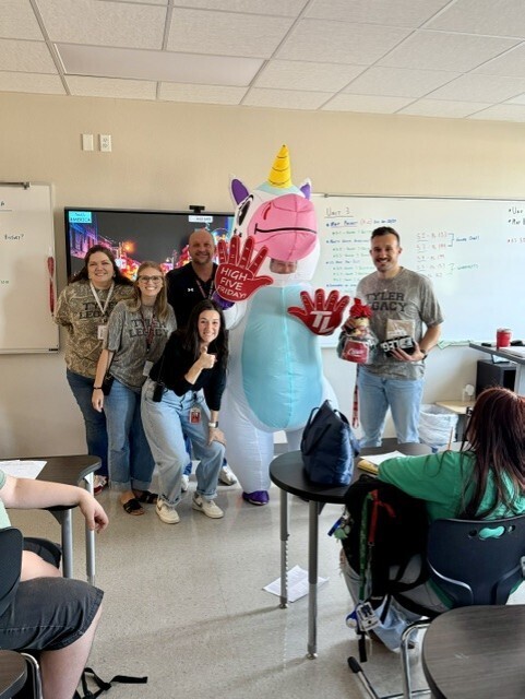 Group of people, one in unicorn costume, in a classroom. Some sit, some stand, and one holds an object.