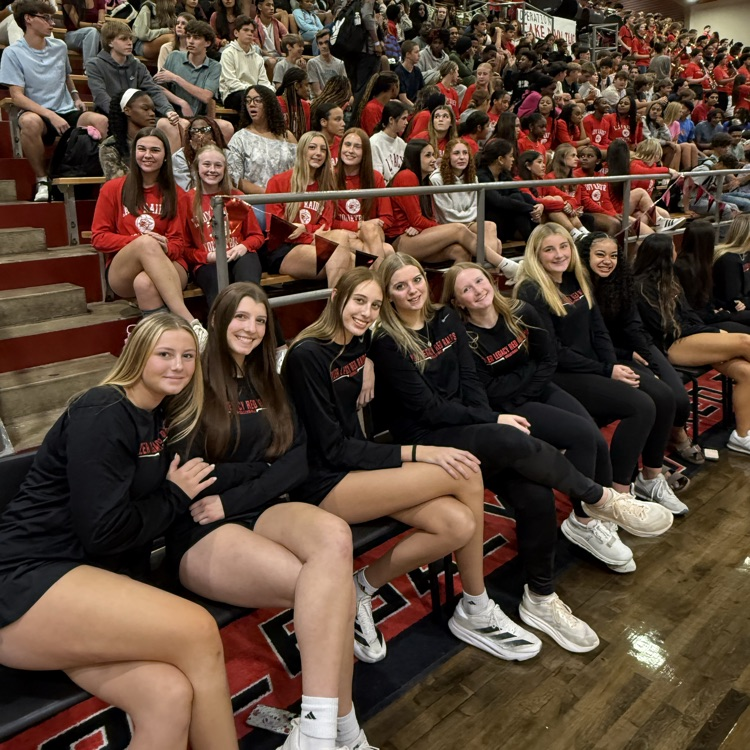 A group of young women wearing matching uniforms sit on bleachers in a gym. Behind them, a crowd of students fills the stands.
