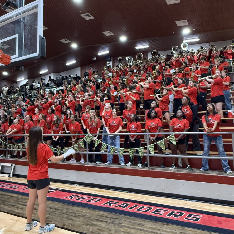 A group of people in red shirts stand in bleachers. A person leads in front wearing red and black.