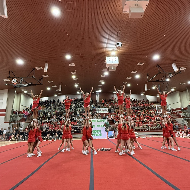 A cheerleading team in red uniforms performs a pyramid stunt in a gymnasium, with spectators seated on bleachers.
