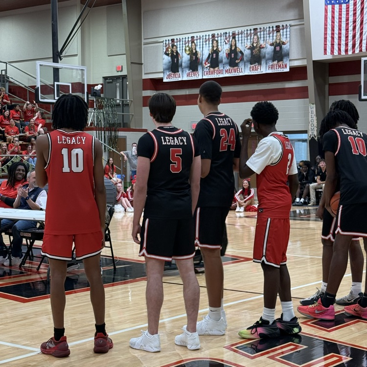 Several basketball players stand in a row on a court. A crowd sits behind them.