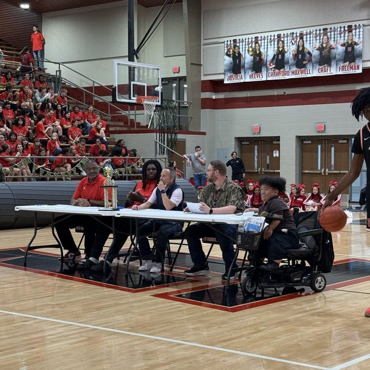 People sit at tables inside a gymnasium. A basketball player holds a ball. Audience members sit on bleachers.